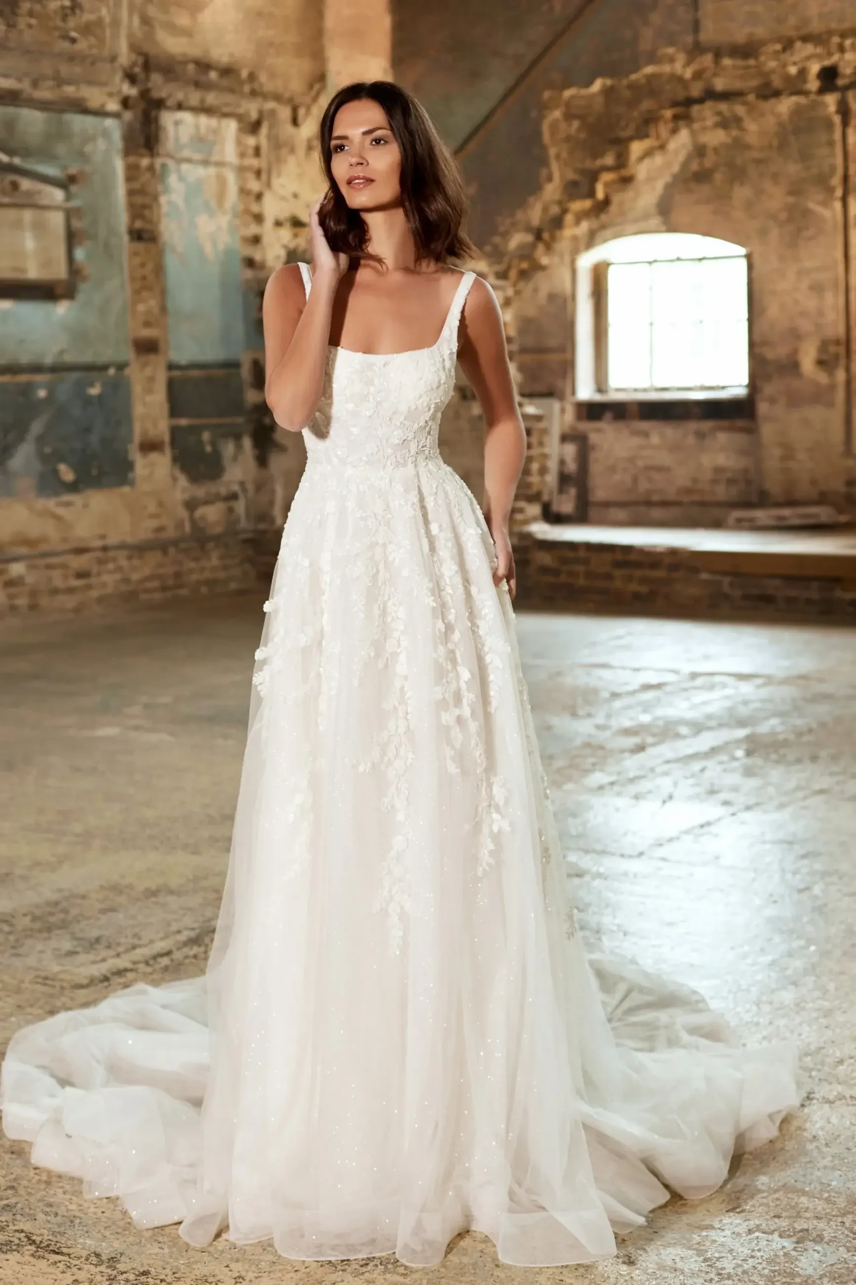 Model is wearing a gown featuring a modern square neckline, gentle A-line silhouette, beaded lace overlay, and a dramatic train.