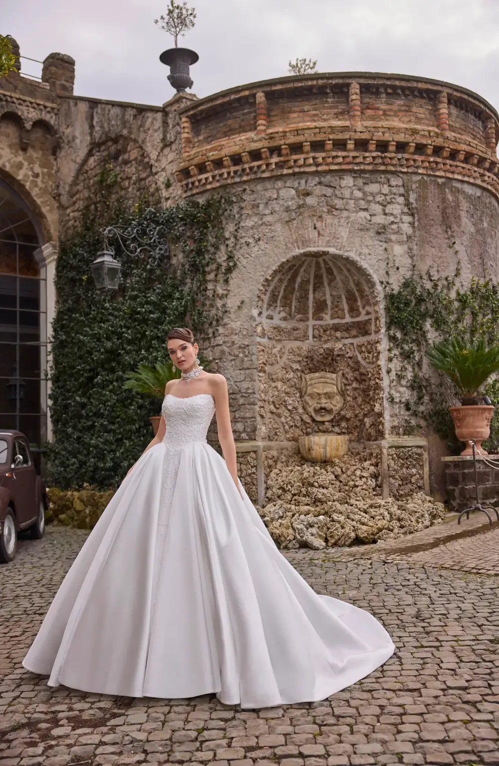 A bride in a white wedding dress stands in front of a rustic stone wall and fountain, surrounded by greenery.