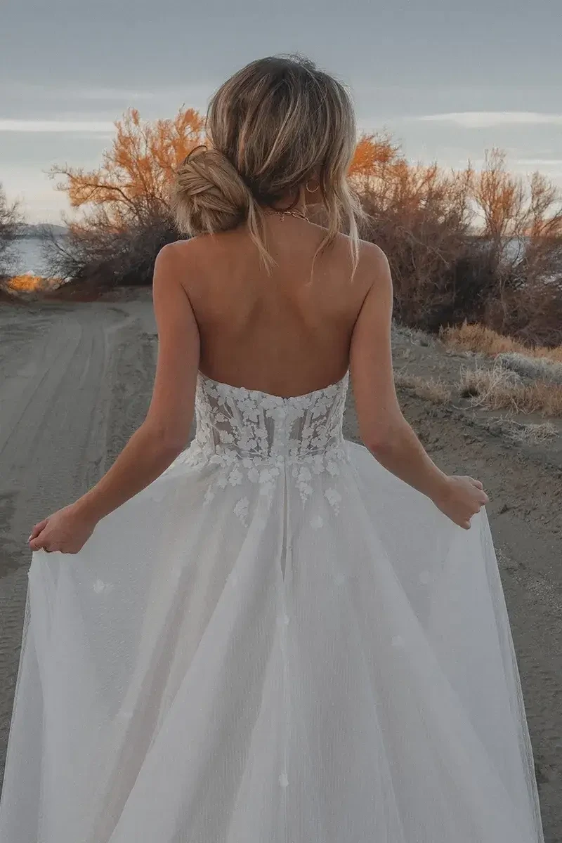 Model is wearing a strapless sweetheart neckline gown with a lace bodice, tulle skirt adorned with floral appliques, and a subtle slit.