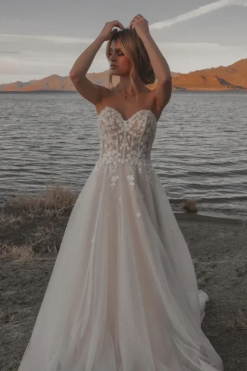 Model is wearing a strapless sweetheart neckline gown with a lace bodice, tulle skirt adorned with floral appliques, and a subtle slit.