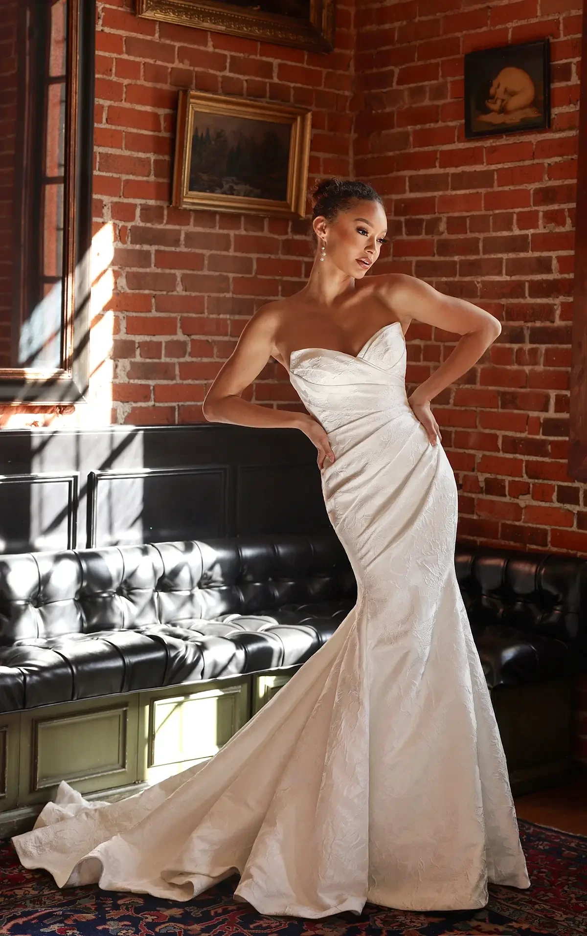 Model is wearing a strapless satin column gown with a draped sweetheart neckline, ruched bodice, and detachable pleated overskirt, finished with vintage-inspired button details.