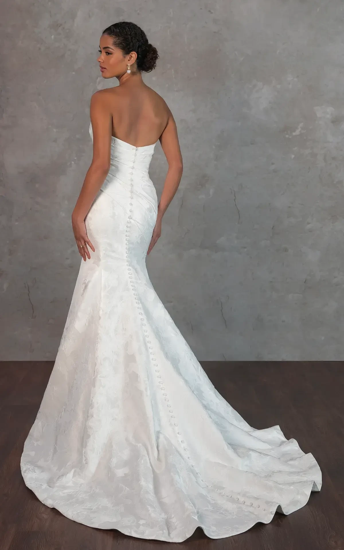 Model is wearing a strapless satin column gown with a draped sweetheart neckline, ruched bodice, and detachable pleated overskirt, finished with vintage-inspired button details.