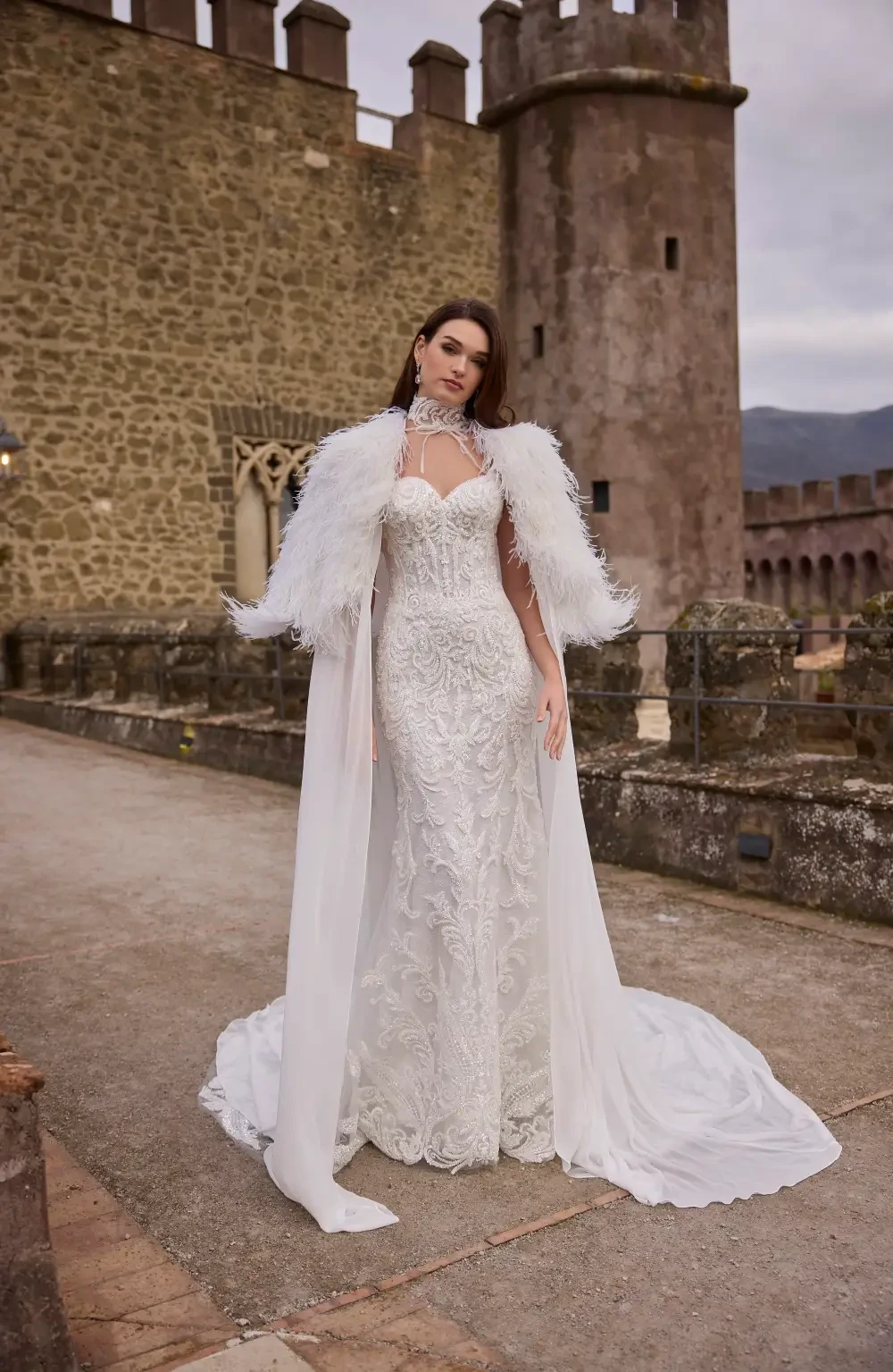 A model in an elegant white wedding dress with intricate details and a feathered cape, standing outdoors near a castle-like structure.