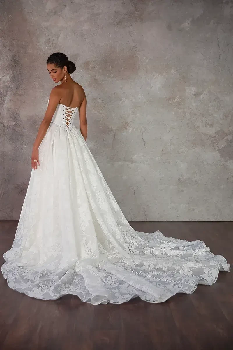 Model is wearing a strapless scoop neckline ballgown with a Basque waist, textured organza skirt with pockets, and a lace-up back.