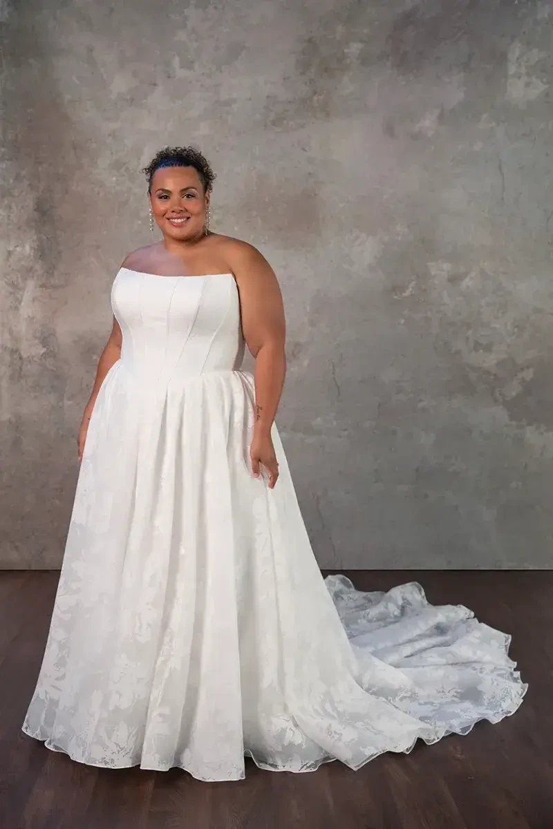Model is wearing a strapless scoop neckline ballgown with a Basque waist, textured organza skirt with pockets, and a lace-up back.
