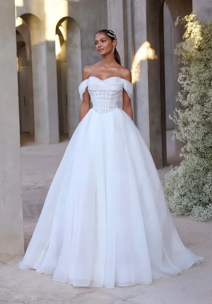 A model wearing a stunning off-shoulder white wedding gown with a fitted bodice and flowing skirt, set in an elegant architectural environment.