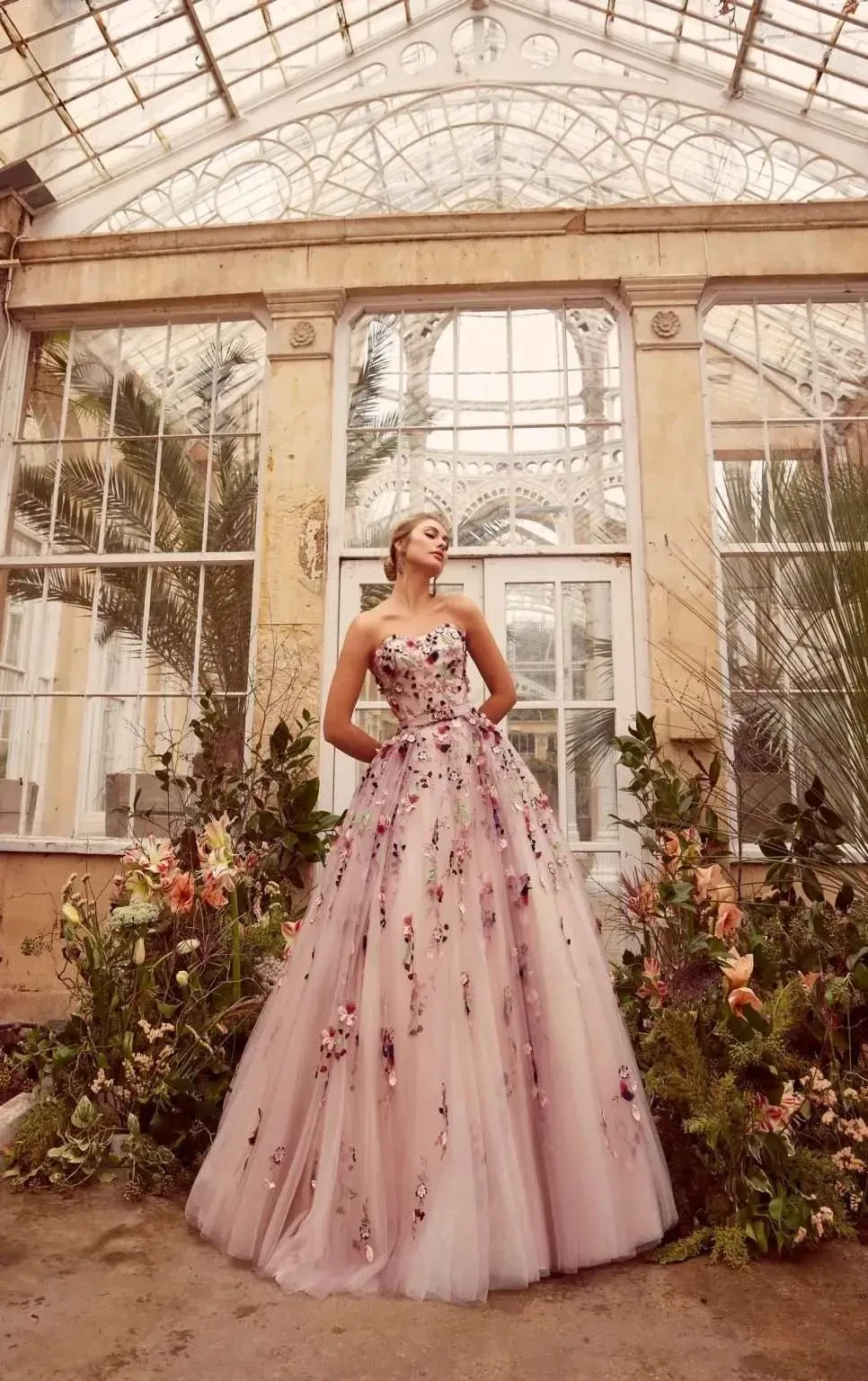 A woman wearing a floral ball gown stands elegantly in a greenhouse filled with plants and flowers.