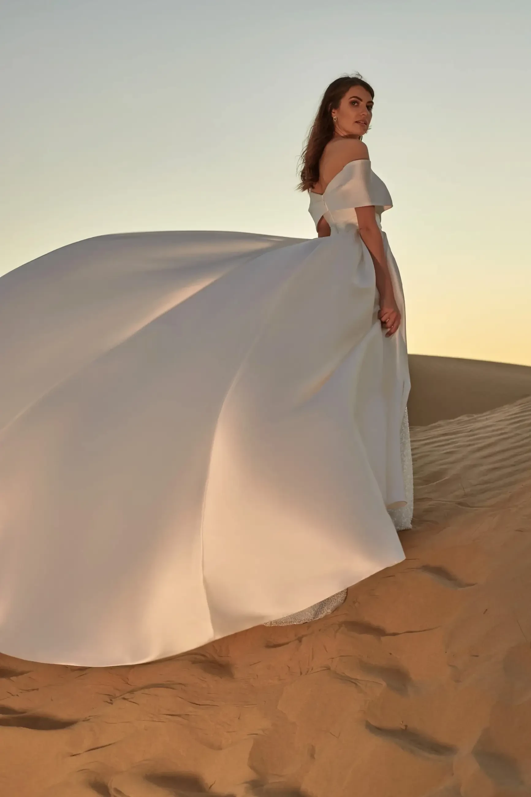 Model is wearing a fitted satin gown with a ruched bodice, bardot neckline, and a shimmering beaded skirt, complete with a detachable satin cathedral train