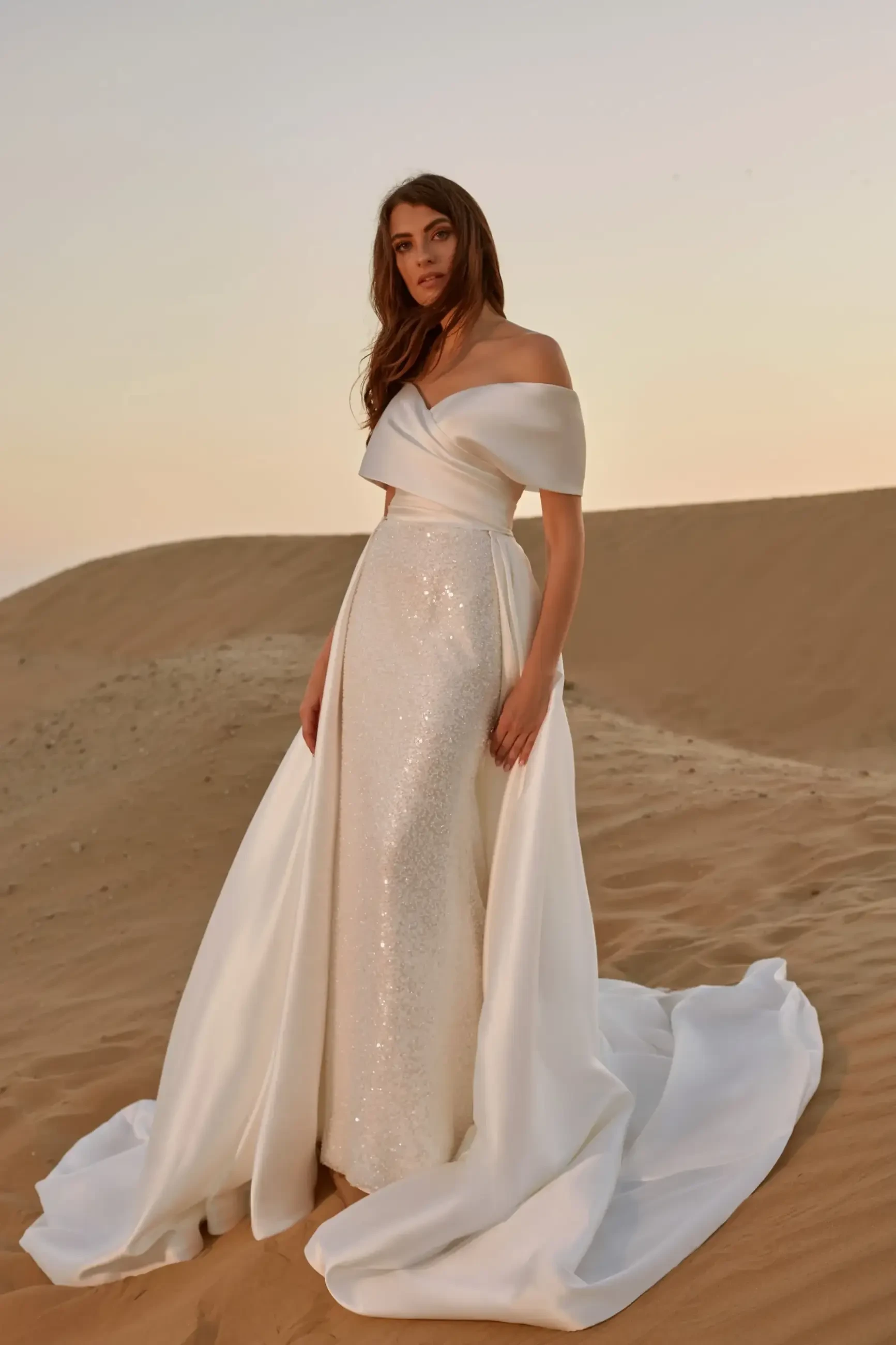 Model is wearing a fitted satin gown with a ruched bodice, bardot neckline, and a shimmering beaded skirt, complete with a detachable satin cathedral train