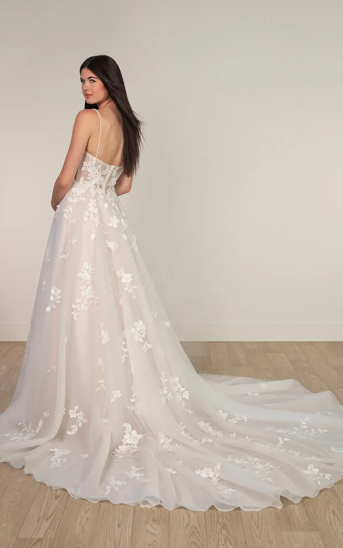 A woman in a white wedding gown stands indoors on a wooden floor. The dress has floral lace details and a long train, exuding elegance and grace.