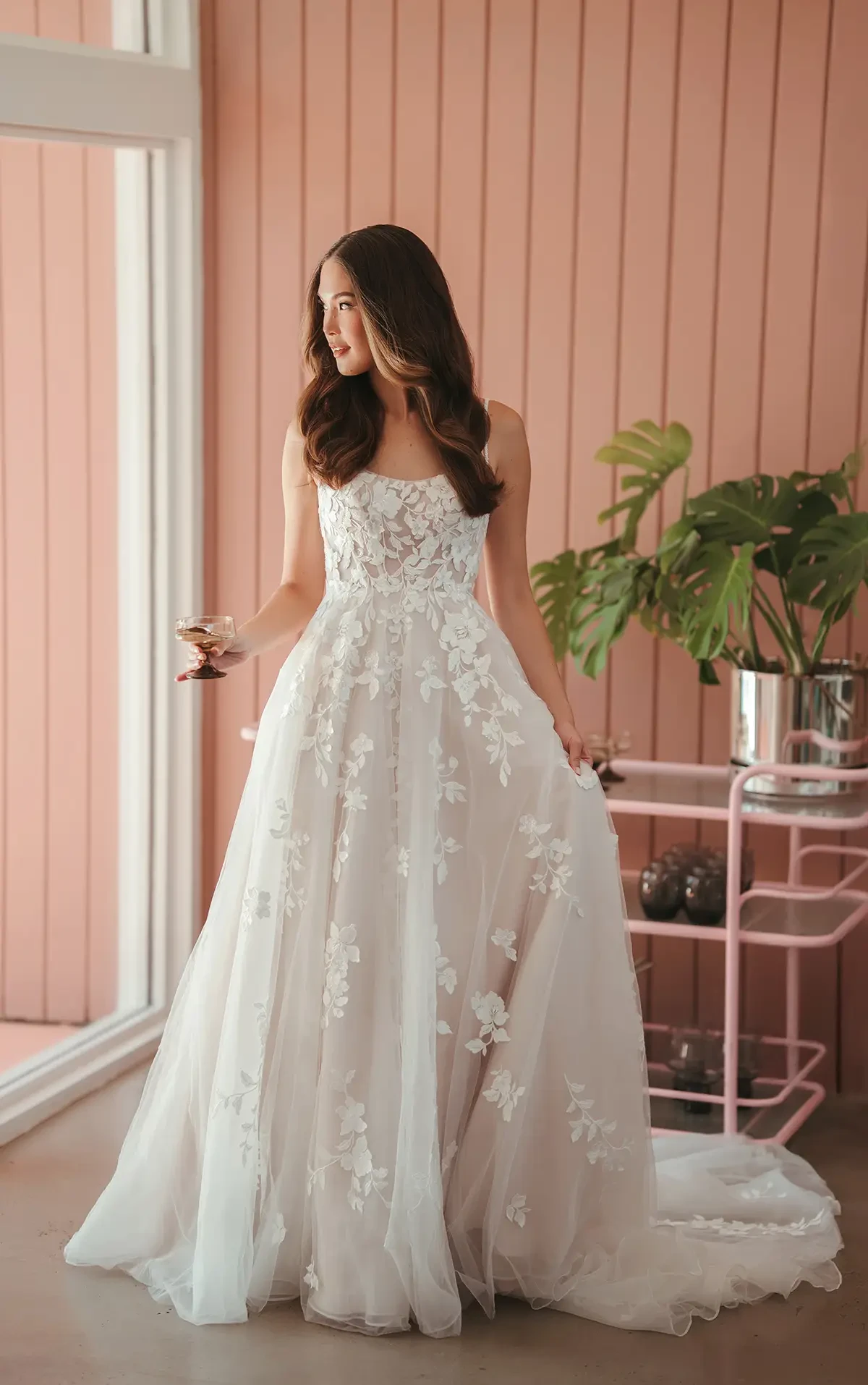 A woman in a white floral lace wedding gown holds a glass, standing by a pink paneled wall with a large plant. She gazes to the side, looking serene.