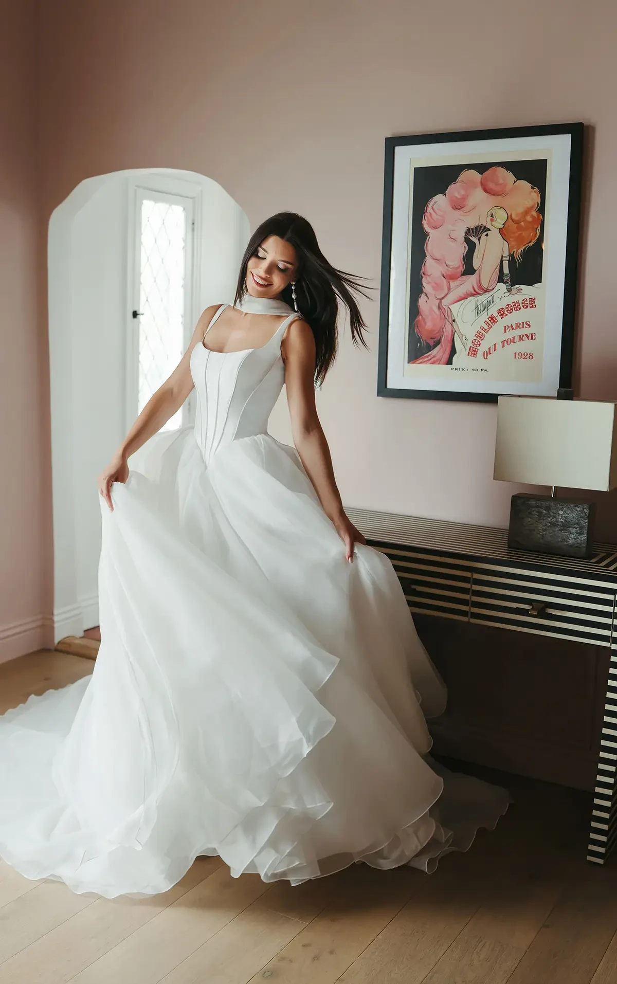 Woman in a flowing white wedding dress stands gracefully in a softly lit room, smiling. Art deco poster on the wall adds a vintage touch.