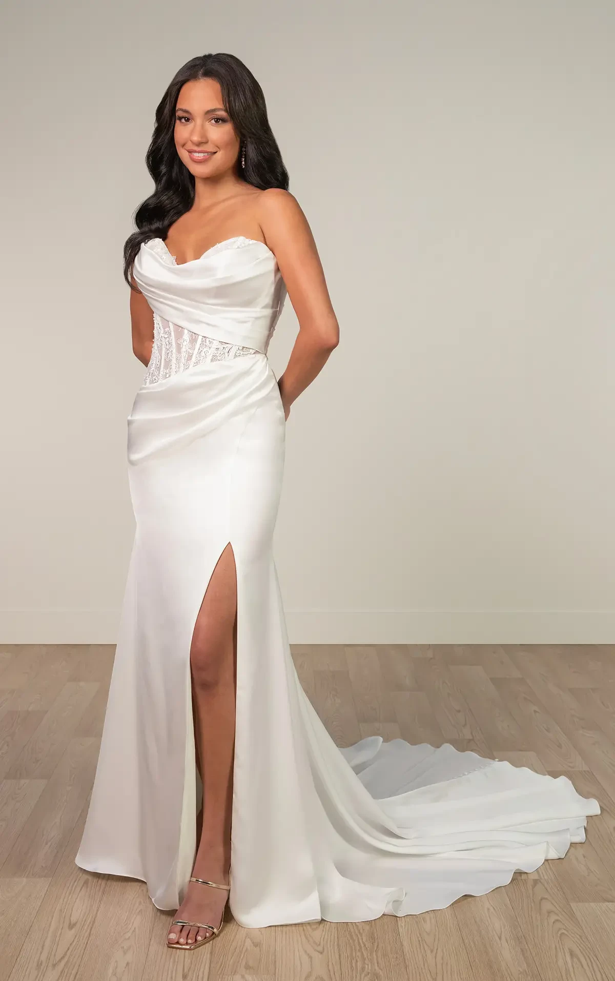 A woman in a sleek, strapless white gown with a lace corset and thigh-high slit stands on a wooden floor, smiling with an elegant and confident pose.