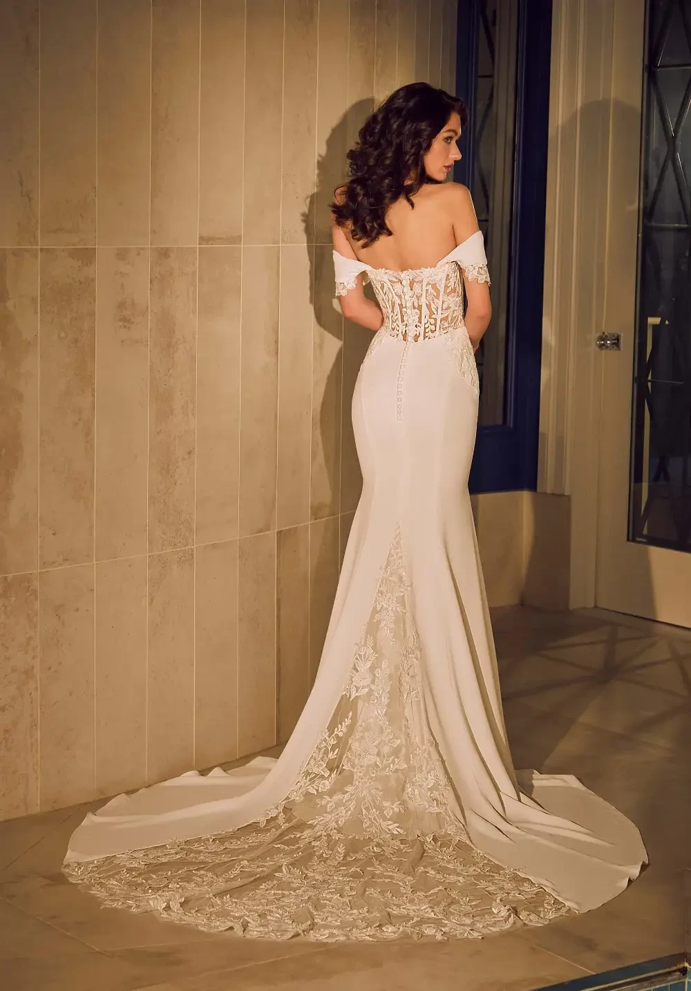 A woman in a fitted white wedding dress with lace details, standing with her back to the camera against a tiled wall.