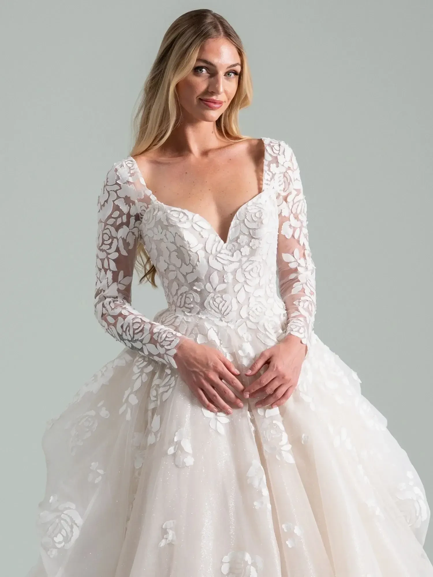 A woman in a white wedding dress with long sheer sleeves adorned with floral embroidery, standing gracefully and smiling.