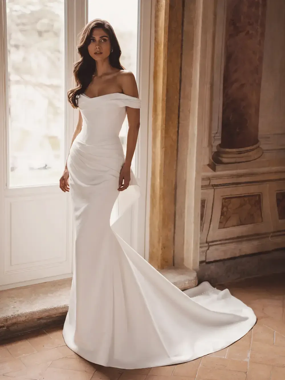 A woman wearing a sleek, off-the-shoulder white gown standing gracefully near a large window with soft natural light.