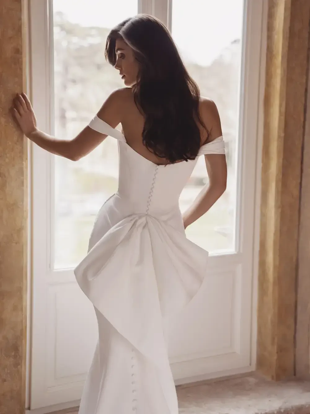 A woman in a white off-the-shoulder dress with a large bow at the back, standing by a window.