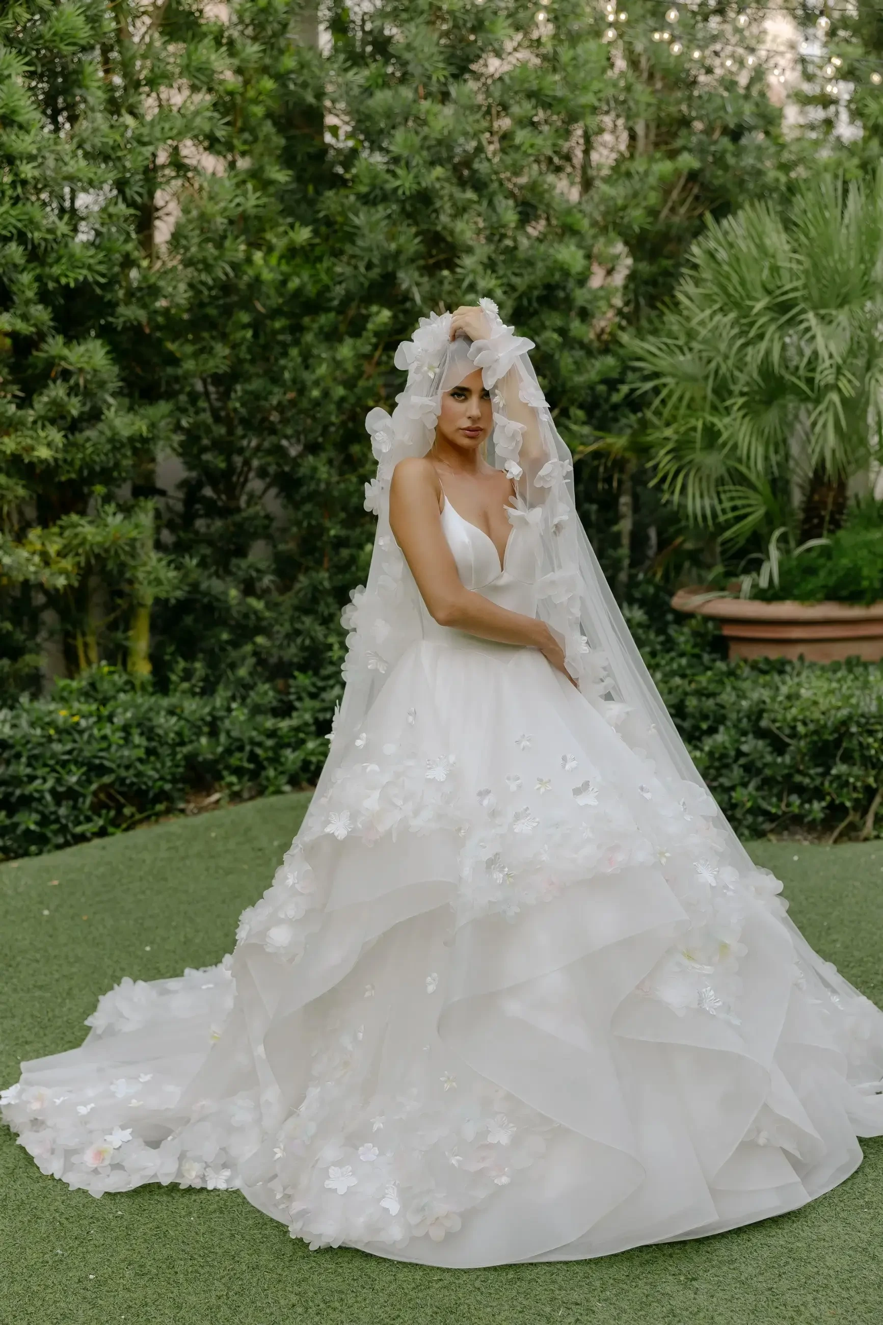 A bride wearing a layered white wedding dress with floral embellishments and a long veil, standing in a garden setting surrounded by greenery.
