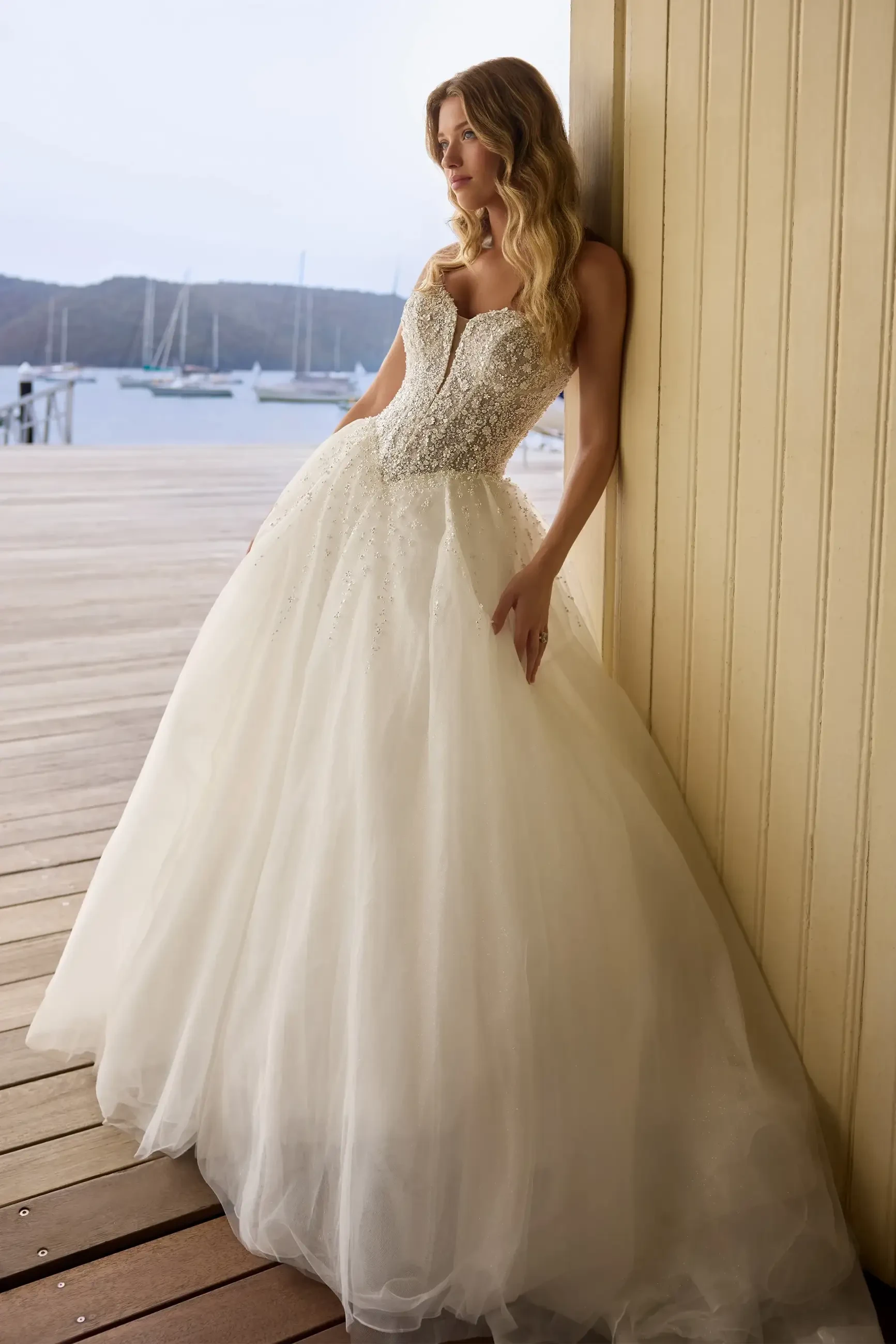 A woman in a stunning strapless wedding gown with a fitted bodice and a full, flowing skirt stands against a wooden wall by the water.