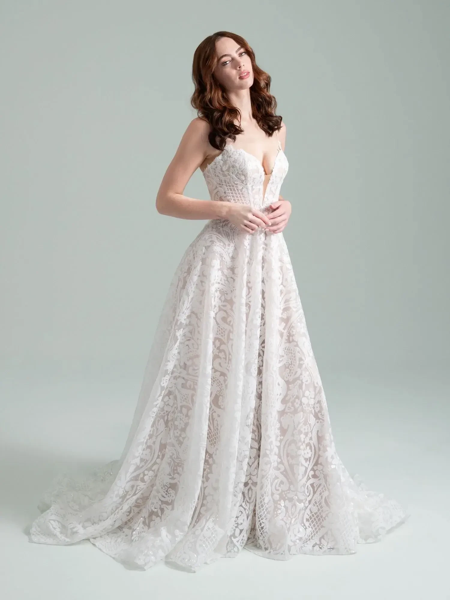 A woman stands gracefully in a flowing white lace gown with a deep neckline and intricate patterns. Her hair is styled in soft curls, and she has a joyful expression. The background is neutral to emphasize the dress.