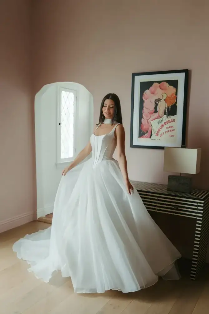 A woman wearing a white bridal gown stands gracefully in a room with pink walls, next to a striped table and framed artwork.